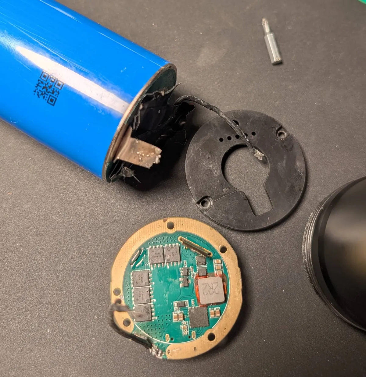 A blue cylindrical battery with a metal tab at one end, next to the flashlight's black front cover and its round driver board with MOSFETs and an inductor.