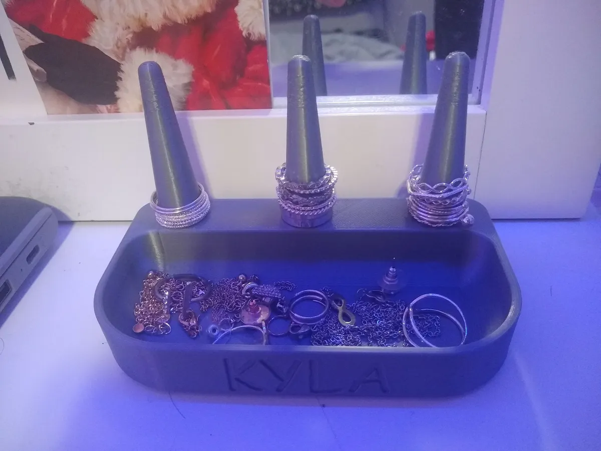 A charcoal printed jewelry organizer with three ring cones at the back, a shallow front tray holding jewelry, and KYLA recessed into the front face.