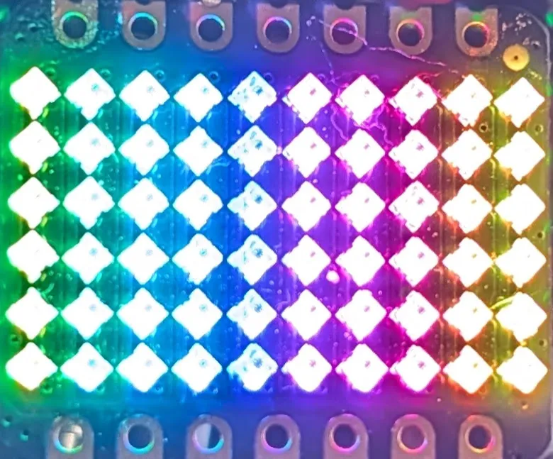 Close-up photo of a compact 6-by-10 RGB LED matrix on a dark PCB running a left-to-right rainbow gradient, with XIAO-style header pads along the top and bottom edges.